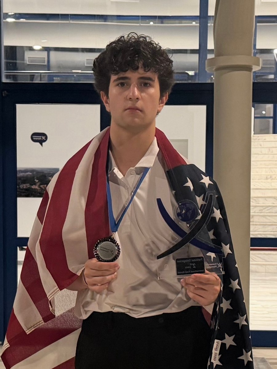Devin Acar wrapped in an American flag, holding a medal and a trophy from the 2026 International Economics Olympiad