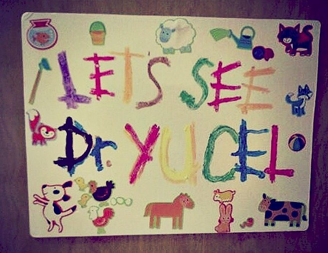 A hand-painted sign made by children reading Let's See Dr. Yucel, decorated with animals and toys, hanging in the office.